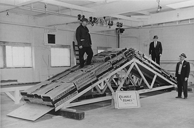 Black and white photograph of a set of roof trusses sitting on the floor with loads and two men standing on top and one man in front of the trusses