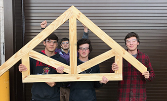 Four students wearing eye protection hold up a roof truss example that they built