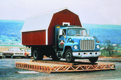 A blue truck with a white and red shed in the truck bed is on top of a set of floor trusses that are on the ground